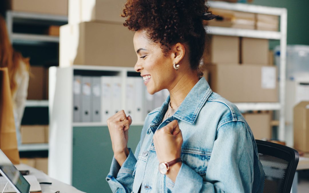 A smiling local business owner celebrates a successful marketing milestone while working on a laptop in a modern office with shelves, boxes, and office supplies in the background.